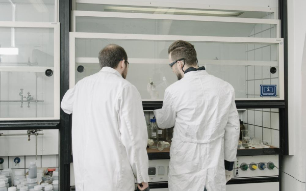 Zwei Männer im Kittel stehen vor einem Abzug im Chemielabor und betrachten eine Probe