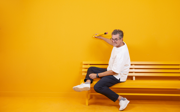Das Bild zeigt Prof. Gunnar Spellmeyer kreativ auf einer Bank vor gelber Wand