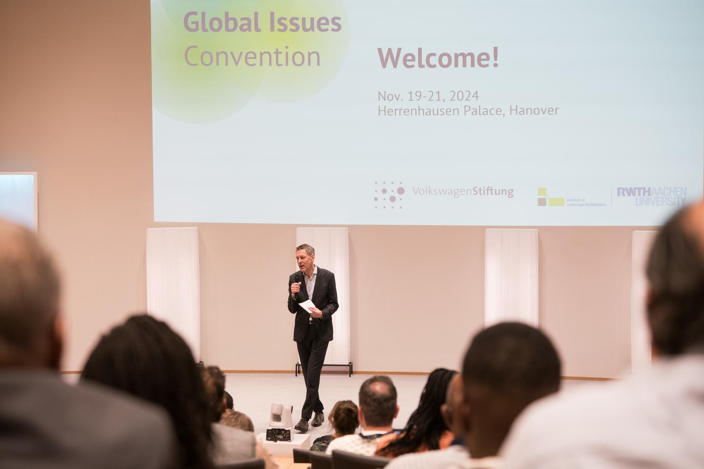 Begrüßung durch einen Mann auf der Bühne, dahinter "Global Issues" und "Welcome!" and die Wand projeziert