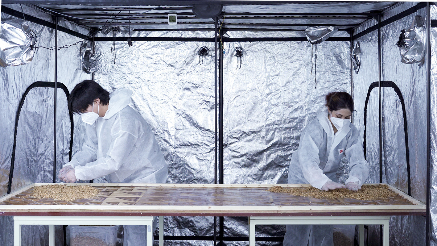Zwei Personen in Ganzkörperanzügen stehen an einem Arbeitstisch in einer Laborumgebung, die durch silberne Folie von der Umgebung abgetrennt ist. Sie arbeiten an einer Form auf dem Tisch.