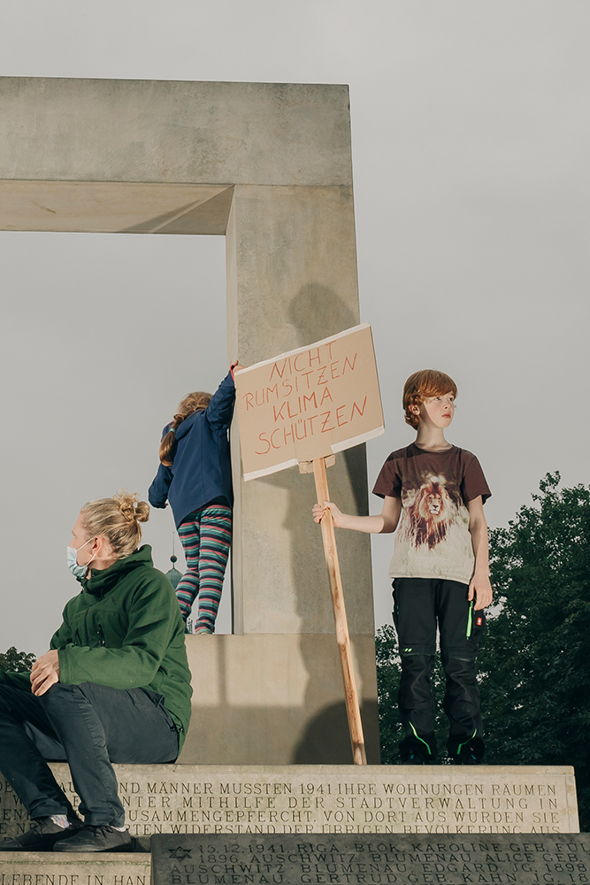 Ein Junge steht auf einem Denkmal und trägt ein Schilf mit der Aufschrift "Nicht rumsitzen, Klima schützen"