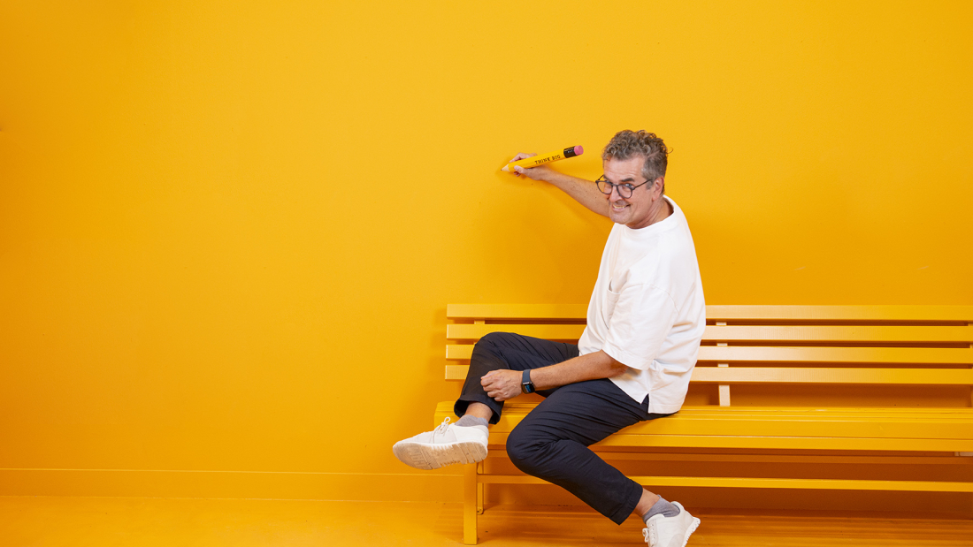 Das Bild zeigt Gunnar Spellmeyer mit einem kreativen Stift auf einer Bank vor einer gelben Wand sitzend und schreiben